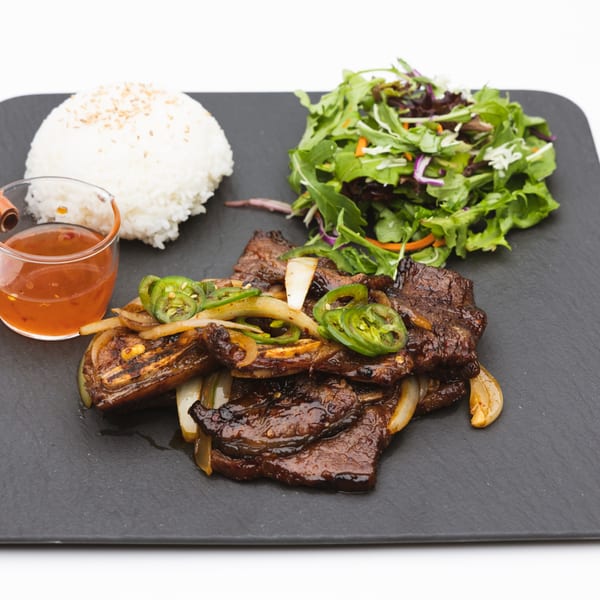 Korean BBQ beef with onions, jalapeños, rice, salad, and a side of sauce on a black plate.