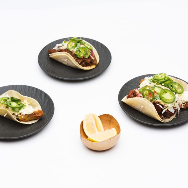 Three Korean tacos with meat, cabbage, and sliced jalapeños on black plates, with lemon wedges.